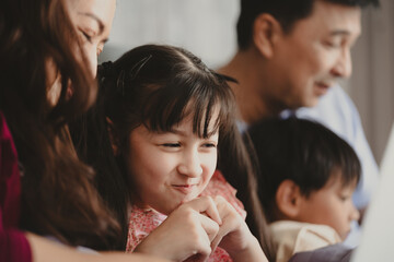 Happy Asian family lying on bed in bedroom with smile, father and mother with daughter and son, lovely parents using laptop computer for education and spent quality time together, laughing together