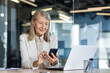 © Liubomir - Senior mature experienced woman at workplace inside office, business woman happy smiling using phone sitting with laptop, gray haired woman boss using app on smartphone typing message.