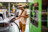 Fototapeta Sypialnia - Man in hat plugs a cable in electric vehicle, while standing with phone on a public charging station outdoors. Concept of travel by electric car and green energy for driving
