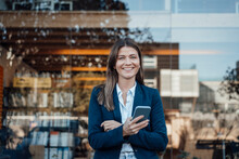 Smiling Businesswoman Holding Smart Phone In Front Of Glass