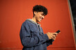 © mimagephotos - Side of smiling young man using mobile phone by red wall