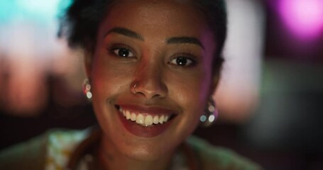 Poster - Happy Creative Young Female Looking Up at Camera and Charmingly Smiling with Colorful Neon Blurry Background. Close Up Portrait of Friendly Black Stylish Woman With a Welcoming Big Smile