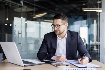 Concentrated young businessman office worker working at workplace with documents. Looks at the laptop monitor and takes notes