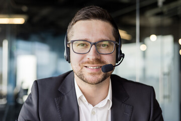 Wall Mural - Close-up photo of a young male teacher wearing a headset and suit sitting in front of the camera and teaching remotely via online video call