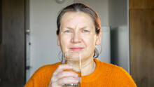 Mature Woman In Cozy Sweater Drinks Glass Of Water In Kitchen. Female With Ponytail Hydrates With Glass Of Water As Part Of Morning Routine