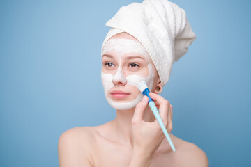 Young woman with face mask and towel on her head holding fresh cucumber slices. Happy girl taking care of her facial skin on blue background.