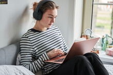 Young teen woman wearing headphones sitting on bed and looking at laptop