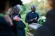 © pressmaster - Focus on young pastor with Holy Bible and rosary beads standing in front of coffin with dead person and group of relatives during funeral service