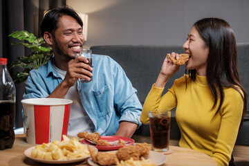 Wall Mural - Asian man and woman couple eating fried chicken happily inside their home celebrating the weekend.