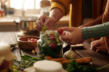 Hands of granddaughter putting fresh tomato into jar with vegetables while standing next to grandmother adding dill to veg assortment