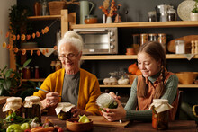 Happy Cute Girl And Her Grandmother Writing Down Pickle Names On Jars While Sitting By Wooden Table After Preparation Of Vegetables For Winter