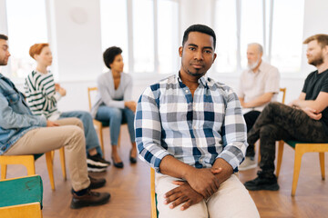 Portrait of confident black male psychologist addiction counsellor looking at camera with serious expression, on background diverse young people sitting in circle share problems supporting each other.