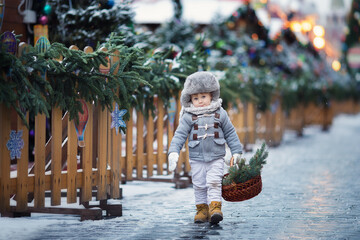 Little boy in grey winter clothes on Christmas fair with basket and pine tree