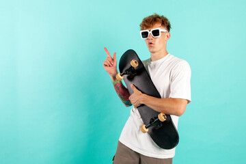 Wall Mural - young cheerful tattooed guy in white t-shirt holds skateboard and smiles on blue isolated background