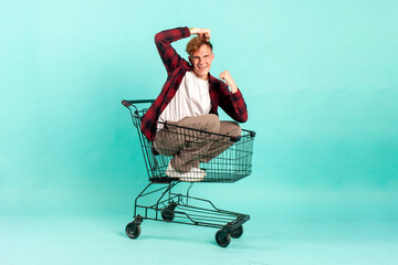 Wall Mural - young cheerful guy winner sits in shopping cart and rejoices in victory on blue background