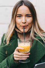 Wall Mural - Close up of blonde young woman looking at camera while drinking orange juice on a straw. Portrait of beautiful young woman with green eyes enjoying her drink.