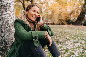 Wall Mural - Young woman using smartphone while sitting on the ground. Smiling attractive woman taking a break in park during the city sightseeing and talking on a phone.