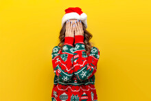 Modest Girl In Christmas Sweater And Santa Claus Hat Covers Her Face With Her Hand And Is Shy On Yellow Background