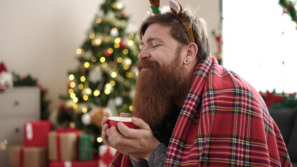 Wall Mural - Young redhead man drinking coffee sitting on sofa by christmas tree at home