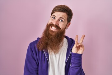 Wall Mural - Caucasian man with long beard standing over pink background smiling with happy face winking at the camera doing victory sign. number two.