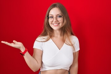 Sticker - Young caucasian woman standing over red background smiling cheerful presenting and pointing with palm of hand looking at the camera.