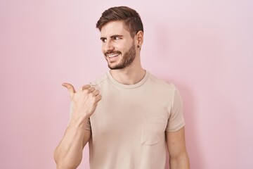 Poster - Hispanic man with beard standing over pink background smiling with happy face looking and pointing to the side with thumb up.