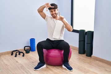 Canvas Print - Hispanic man with beard sitting on pilate balls at yoga room smiling making frame with hands and fingers with happy face. creativity and photography concept.