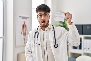 Sticker - Hispanic doctor man with beard holding covid record card scared and amazed with open mouth for surprise, disbelief face