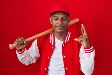 Wall Mural - Young hispanic man playing baseball holding bat gesturing finger crossed smiling with hope and eyes closed. luck and superstitious concept.