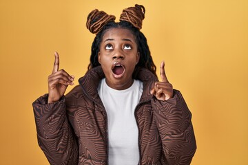 Wall Mural - African woman with braided hair standing over yellow background amazed and surprised looking up and pointing with fingers and raised arms.