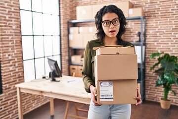 Wall Mural - Hispanic woman with dark hair working at small business ecommerce holding boxes clueless and confused expression. doubt concept.