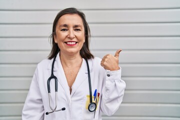 Poster - Middle age hispanic woman wearing doctor uniform and stethoscope pointing thumb up to the side smiling happy with open mouth