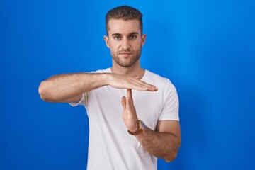 Wall Mural - Young caucasian man standing over blue background doing time out gesture with hands, frustrated and serious face