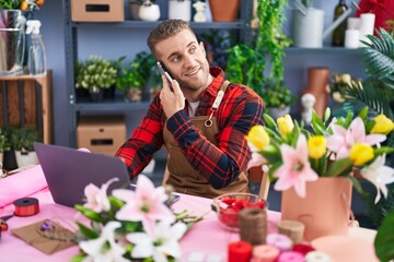 Wall Mural - Young caucasian man florist talking on smartphone using laptop at flower shop