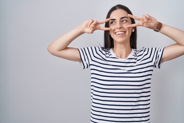 Poster - Young brunette woman wearing striped t shirt doing peace symbol with fingers over face, smiling cheerful showing victory