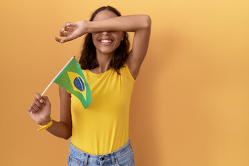 Canvas Print - Young hispanic woman holding brazil flag smiling cheerful playing peek a boo with hands showing face. surprised and exited