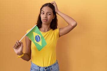 Wall Mural - Young hispanic woman holding brazil flag confuse and wondering about question. uncertain with doubt, thinking with hand on head. pensive concept.