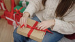 Young beautiful hispanic woman sitting on sofa by christmas tree packing gift at home