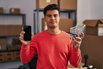 Sticker - Hispanic man working at small business ecommerce holding cart relaxed with serious expression on face. simple and natural looking at the camera.