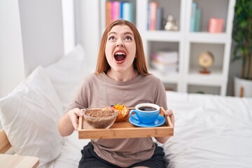 Wall Mural - Redhead woman wearing pajama holding breakfast tray angry and mad screaming frustrated and furious, shouting with anger looking up.