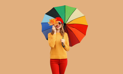 Autumn color style outfit, stylish cheerful young woman holds yellow maple leaves with colorful umbrella wearing red french beret hat, yellow knitted sweater on beige studio background