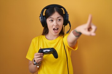 Poster - Chinese young woman playing video game holding controller pointing with finger surprised ahead, open mouth amazed expression, something on the front