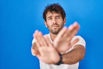 Poster - Hispanic young man standing over blue background rejection expression crossing arms and palms doing negative sign, angry face