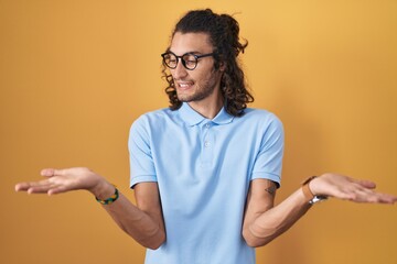 Poster - Young hispanic man standing over yellow background smiling showing both hands open palms, presenting and advertising comparison and balance