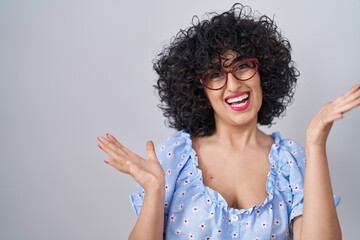 Wall Mural - Young brunette woman with curly hair wearing glasses over isolated background celebrating crazy and amazed for success with arms raised and open eyes screaming excited. winner concept