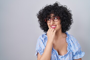 Poster - Young brunette woman with curly hair wearing glasses over isolated background looking stressed and nervous with hands on mouth biting nails. anxiety problem.