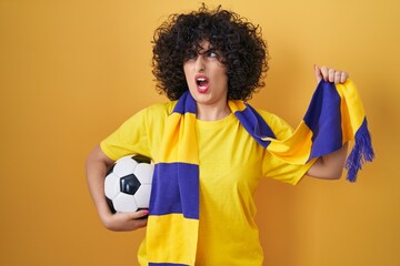 Poster - Young brunette woman with curly hair football hooligan holding ball angry and mad screaming frustrated and furious, shouting with anger. rage and aggressive concept.