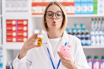 Wall Mural - Young caucasian woman working at pharmacy drugstore holding pills an piggy bank making fish face with mouth and squinting eyes, crazy and comical.