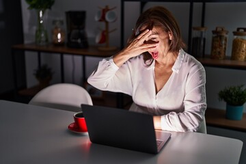 Wall Mural - Middle age hispanic woman using laptop at home at night peeking in shock covering face and eyes with hand, looking through fingers with embarrassed expression.