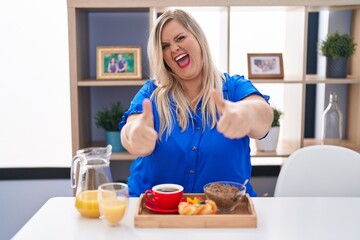 Canvas Print - Caucasian plus size woman eating breakfast at home approving doing positive gesture with hand, thumbs up smiling and happy for success. winner gesture.